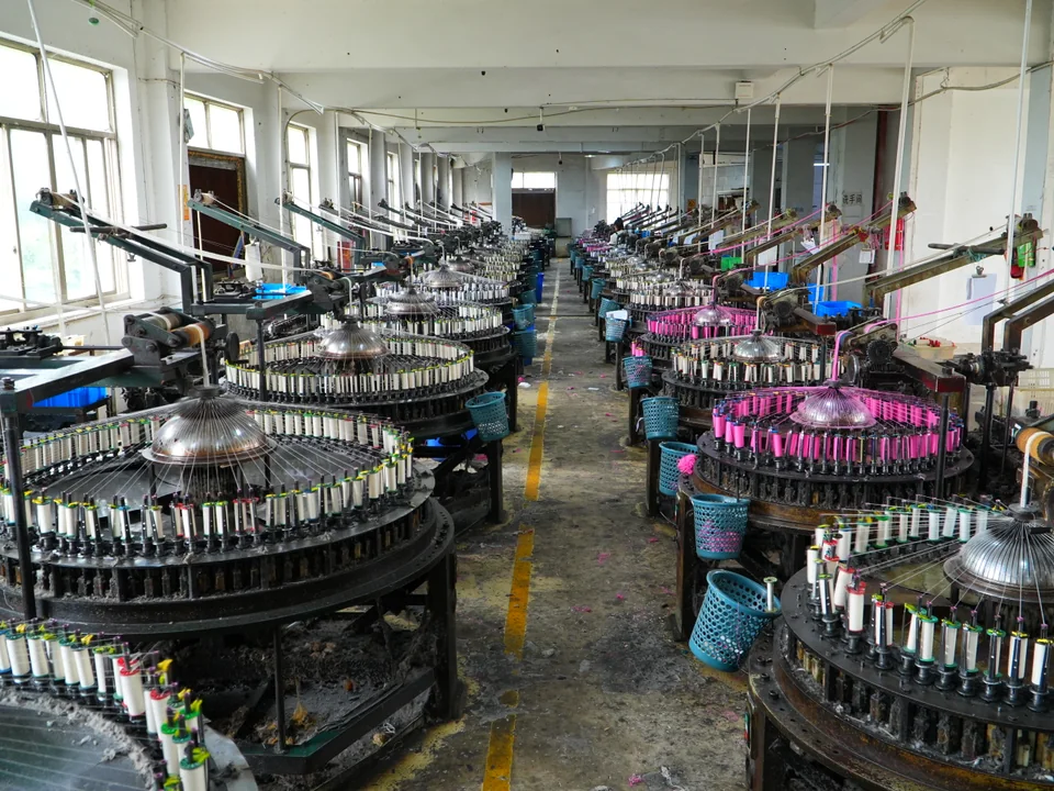 Des rangées de machines textiles circulaires équipées de bobines de fil le long d’une allée lumineuse de l’atelier.