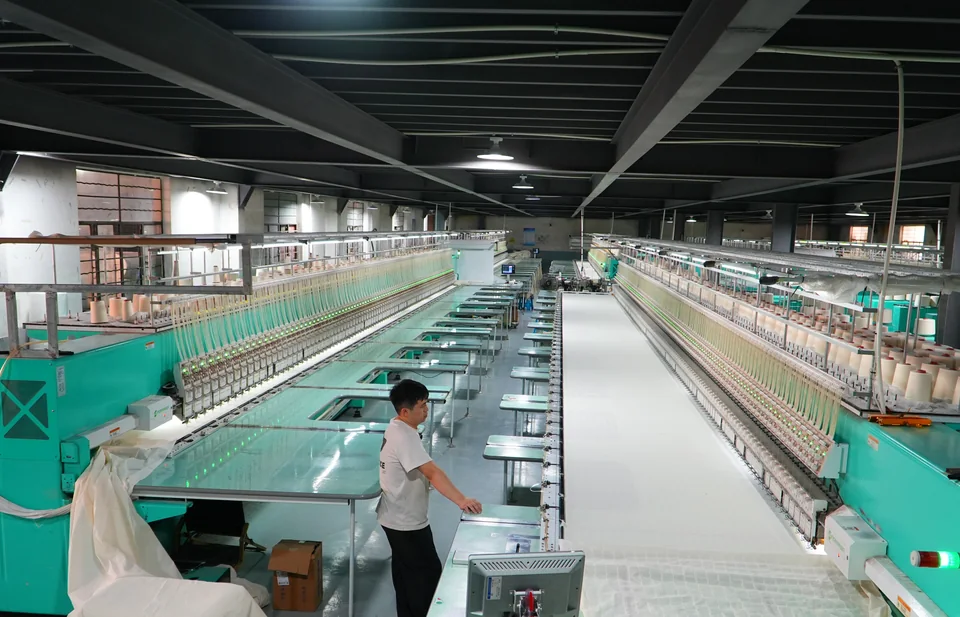 Une personne au travail près de longues machines à broder vertes, avec du tissu blanc tendu le long de la ligne de production.