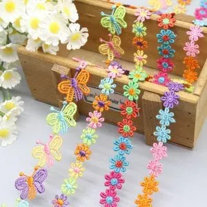 Tiras de encaje hidrosoluble de colores bordadas con mariposas y flores, expuestas sobre una caja de madera con margaritas blancas. Tiras de encaje hidrosoluble de colores bordadas con mariposas y flores, expuestas sobre una caja de madera con margaritas blancas.