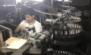 A worker attending a textile machine beside thread spools in a workshop.