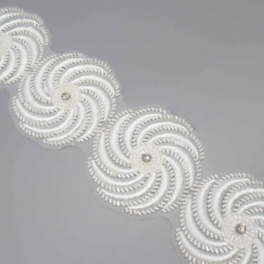 Galón floral blanco en espiral con pétalos bordeados de cuentas dispuesto en diagonal sobre una superficie gris. Galón floral blanco en espiral con pétalos bordeados de cuentas dispuesto en diagonal sobre una superficie gris.