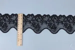 Schwarze Blumenspitzenborte mit Bogenkante neben einem Holzlineal auf einer grauen Fläche.