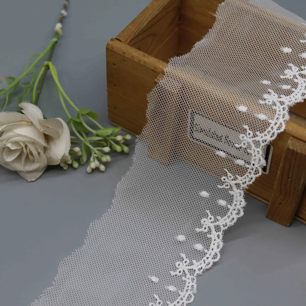 Galon blanc sur résille à points brodés et bordure florale festonnée, drapé sur une boîte en bois. Galon blanc sur résille à points brodés et bordure florale festonnée, drapé sur une boîte en bois.