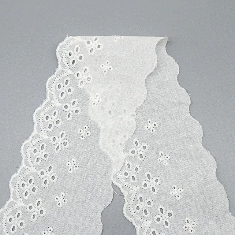 Galon de broderie anglaise blanc disposé en une longue boucle sur une surface grise, laissant voir sa bordure florale festonnée. Galon de broderie anglaise blanc disposé en une longue boucle sur une surface grise, laissant voir sa bordure florale festonnée.