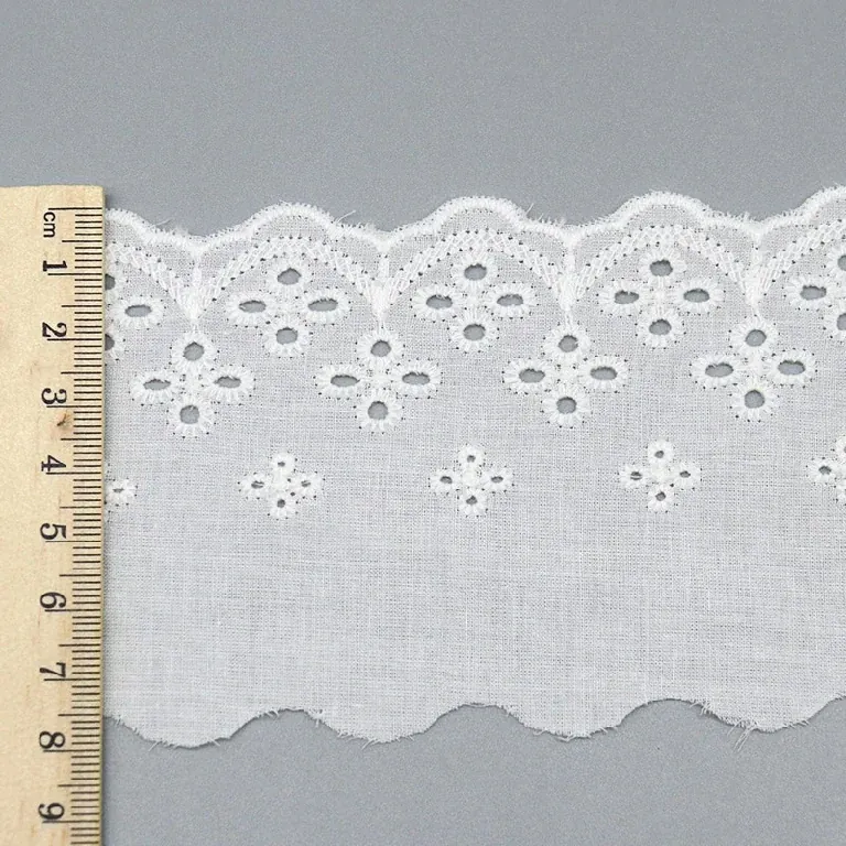 Un mètre ruban posé en travers d’un galon de broderie anglaise blanc à bordure florale festonnée. Un mètre ruban posé en travers d’un galon de broderie anglaise blanc à bordure florale festonnée.
