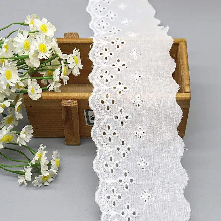 Galon de broderie anglaise blanc à ajours floraux suspendu sur une boîte en bois près de fleurs blanches.