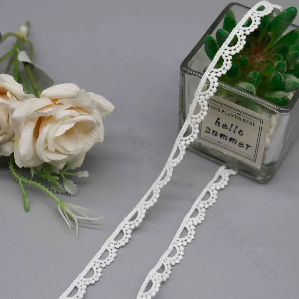 Galon de dentelle blanche à festons en éventail suspendu près d’une jardinière en verre et de roses crème.