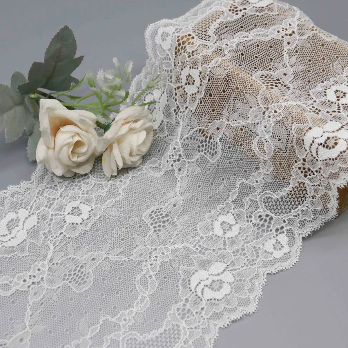 Large dentelle blanche à motifs floraux drapée sur un panier tressé près de roses crème.