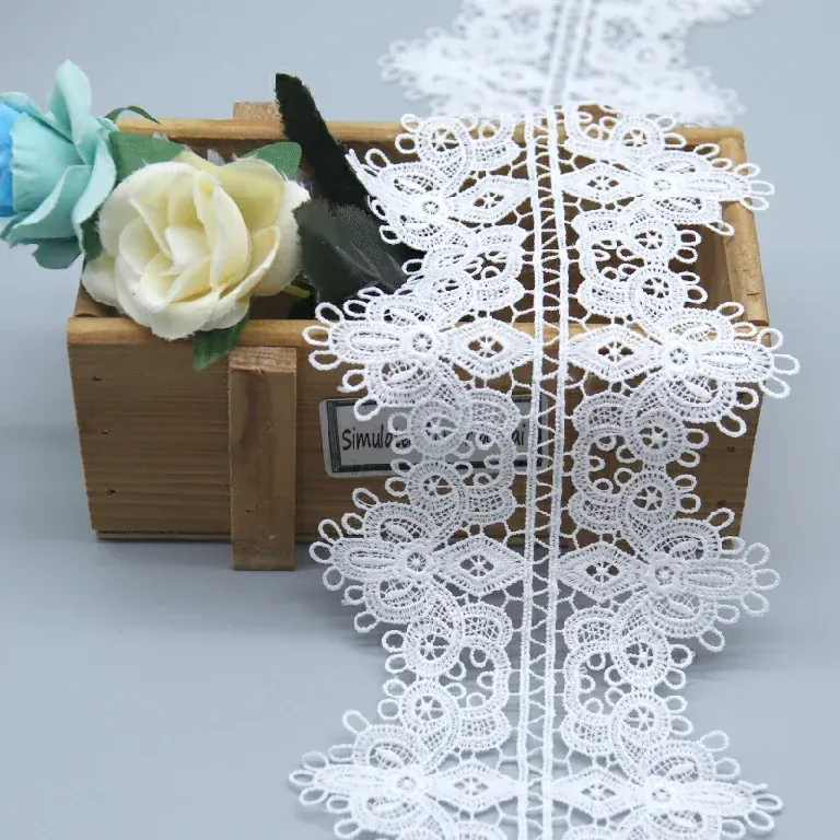 Tira de encaje floral blanco que cae verticalmente sobre una caja de madera junto a flores de tonos claros.