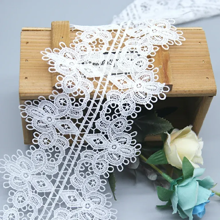 Tira de encaje floral blanco con relieve extendida con caída sobre una caja de madera situada en una superficie gris.