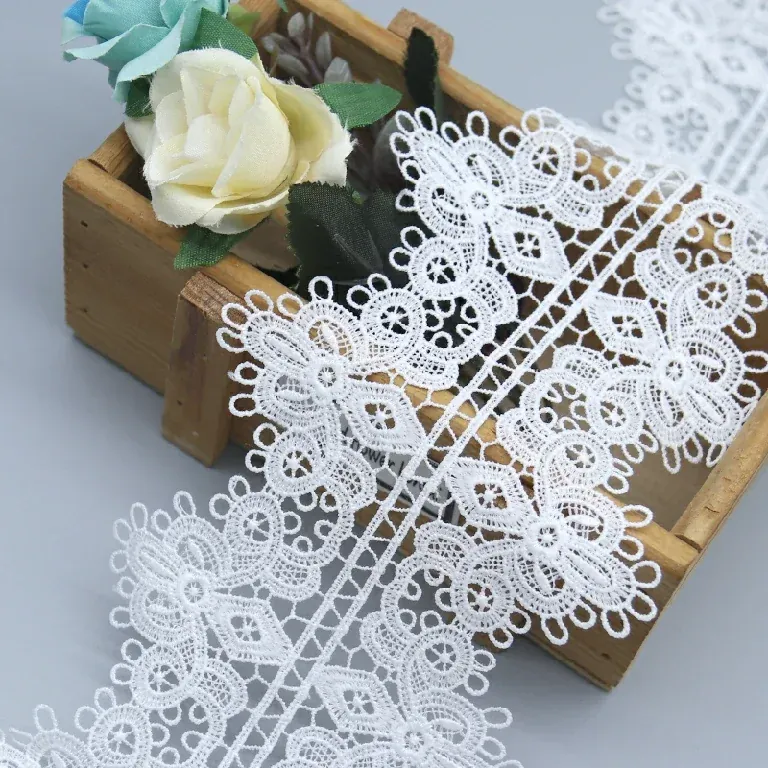 Tira de encaje floral blanco con relieve colocada con caída sobre una caja de madera junto a flores de tonos claros.
