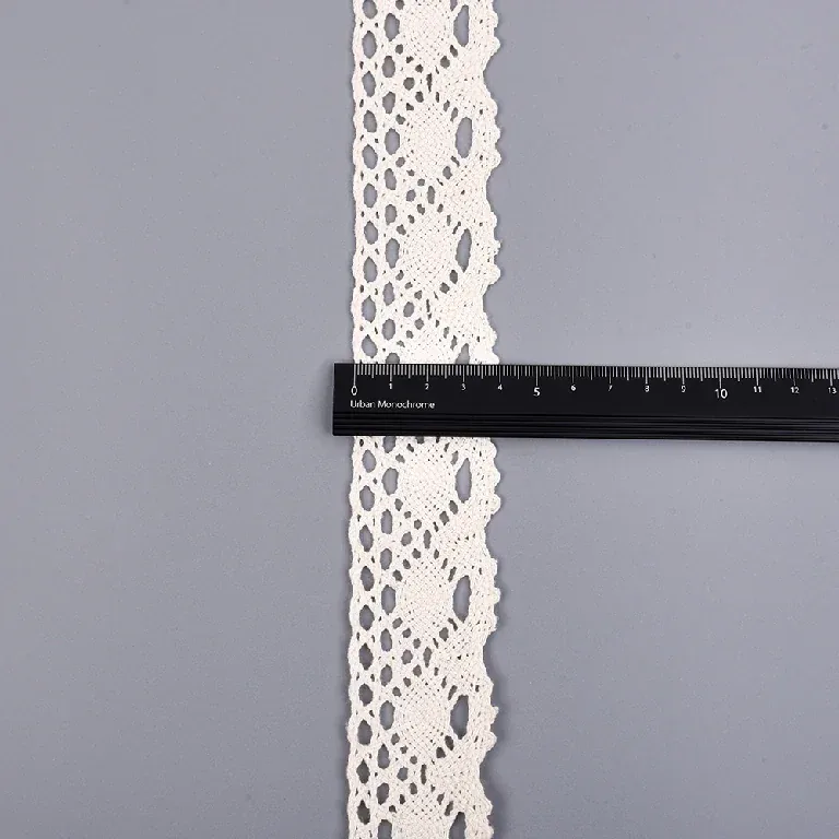 Une règle noire placée en travers d’un galon de dentelle crème à motif d’ovales sur une surface grise.