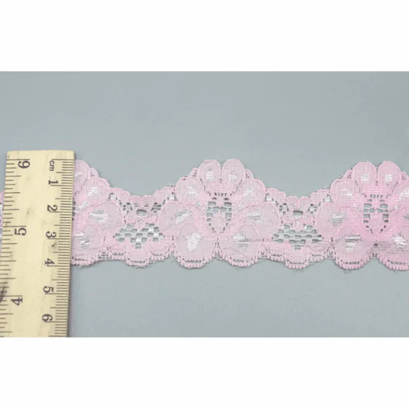 Un mètre ruban posé en travers d’un galon de dentelle rose pâle à motifs floraux et bords festonnés.