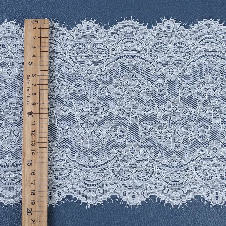 Un mètre ruban posé en travers d’un large galon de dentelle blanche à motifs floraux et bords festonnés et frangés. Un mètre ruban posé en travers d’un large galon de dentelle blanche à motifs floraux et bords festonnés et frangés.