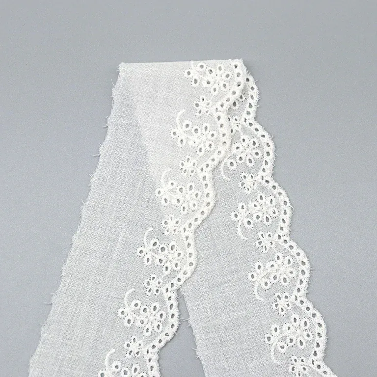 Galon de broderie anglaise blanc disposé en une longue boucle sur une surface grise, laissant voir sa petite bordure florale festonnée.