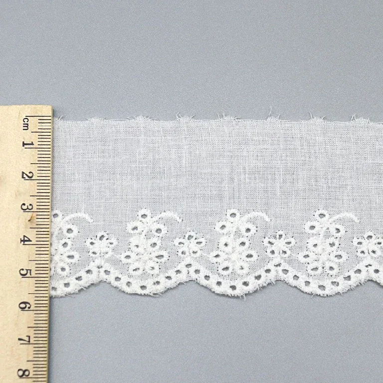 Un mètre ruban posé en travers d’un galon de broderie anglaise blanc à petite bordure florale festonnée.