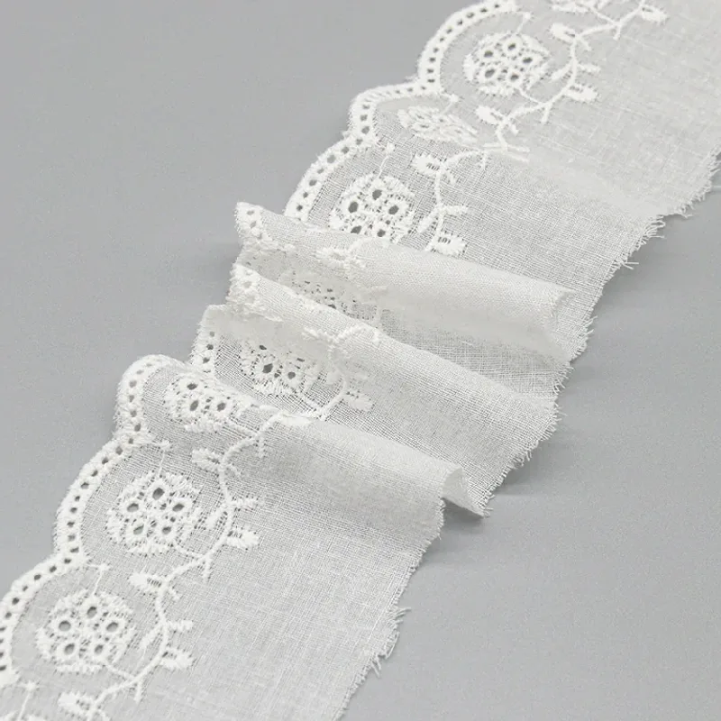 Galon de broderie anglaise blanc à motifs floraux disposé en plis superposés sur une surface grise.