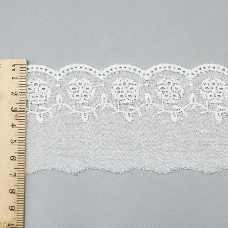 Un mètre ruban posé en travers d’un galon de broderie anglaise blanc à bordure florale arrondie.
