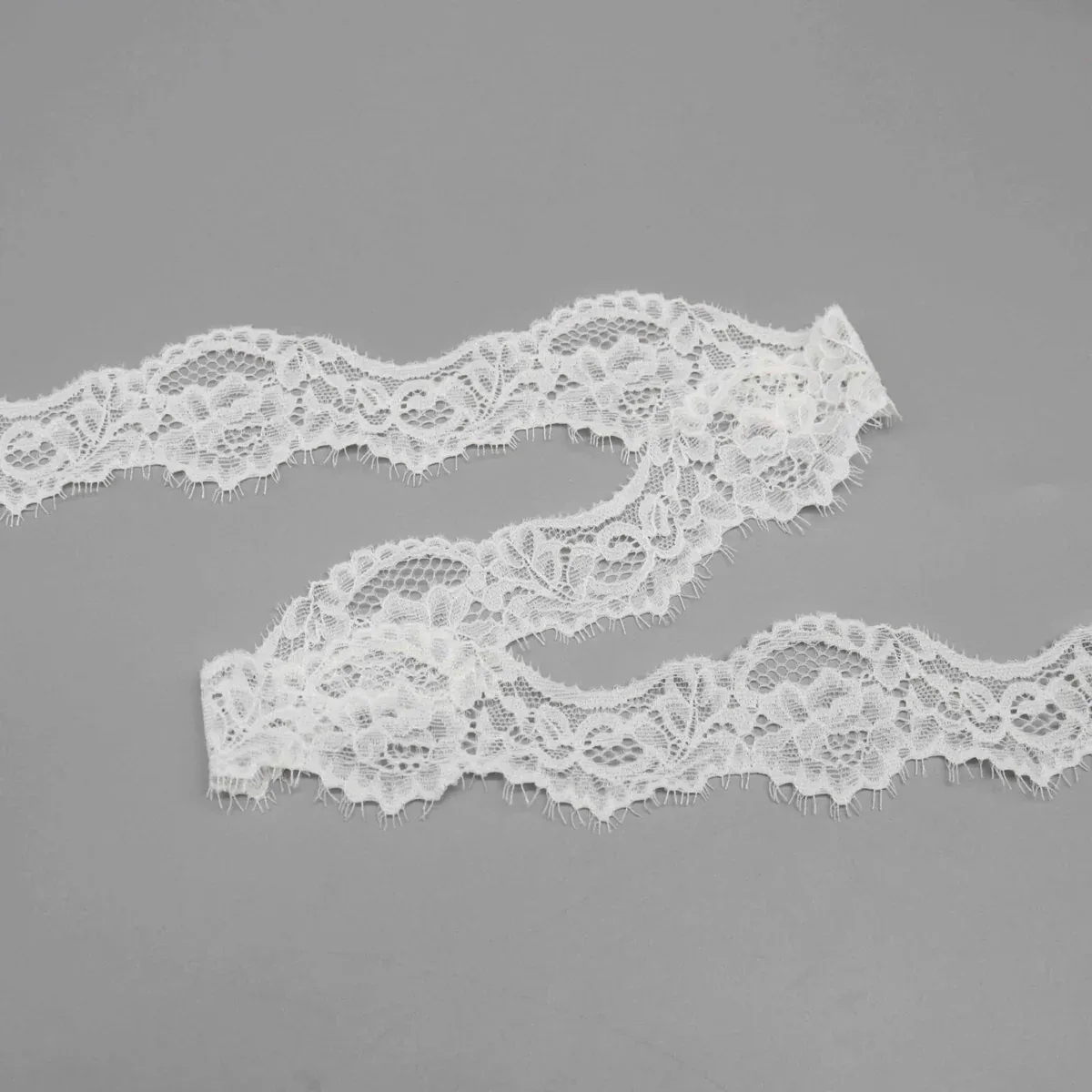 White floral eyelash lace trim arranged in loose waves on a gray surface. White floral eyelash lace trim arranged in loose waves on a gray surface.