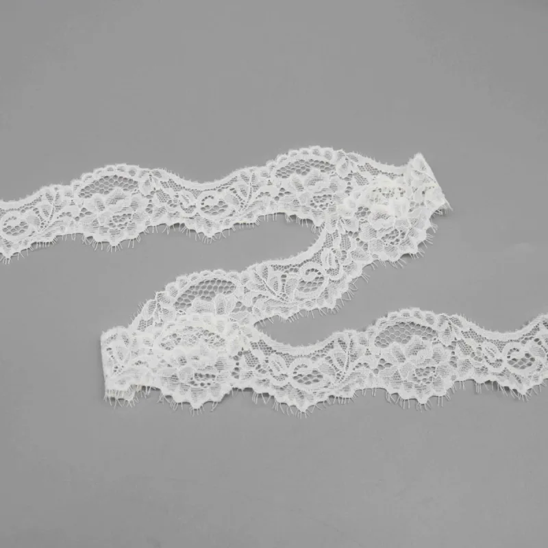 Galon de dentelle blanche à motifs floraux et franges cils disposé en vagues lâches sur une surface grise.