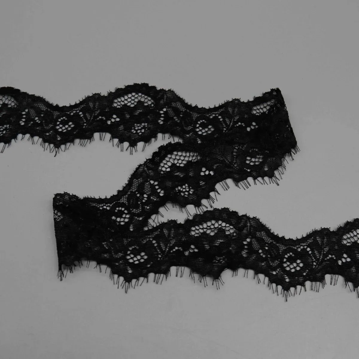 Black floral eyelash lace trim arranged in loose waves on a gray surface. Black floral eyelash lace trim arranged in loose waves on a gray surface.