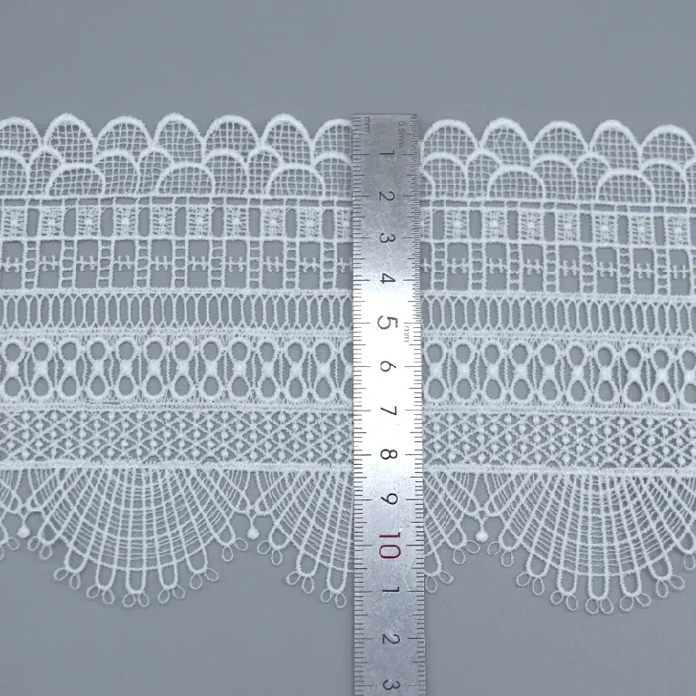 A ruler placed across white geometric lace trim with fan-shaped edges. A ruler placed across white geometric lace trim with fan-shaped edges.