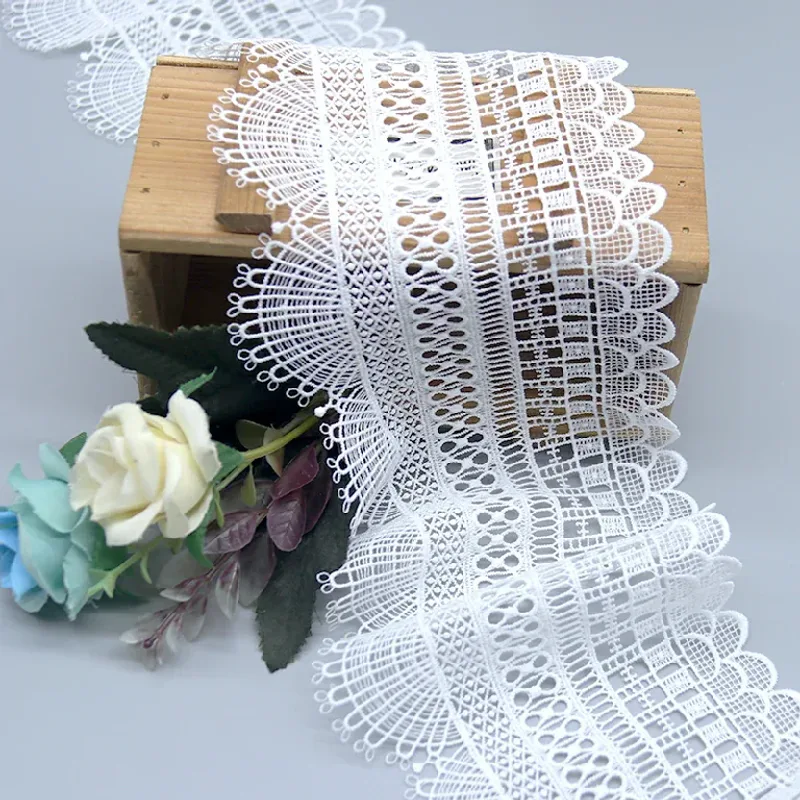 Galon de dentelle blanche aux bords en éventail retombant d’une boîte en bois à côté de fleurs.