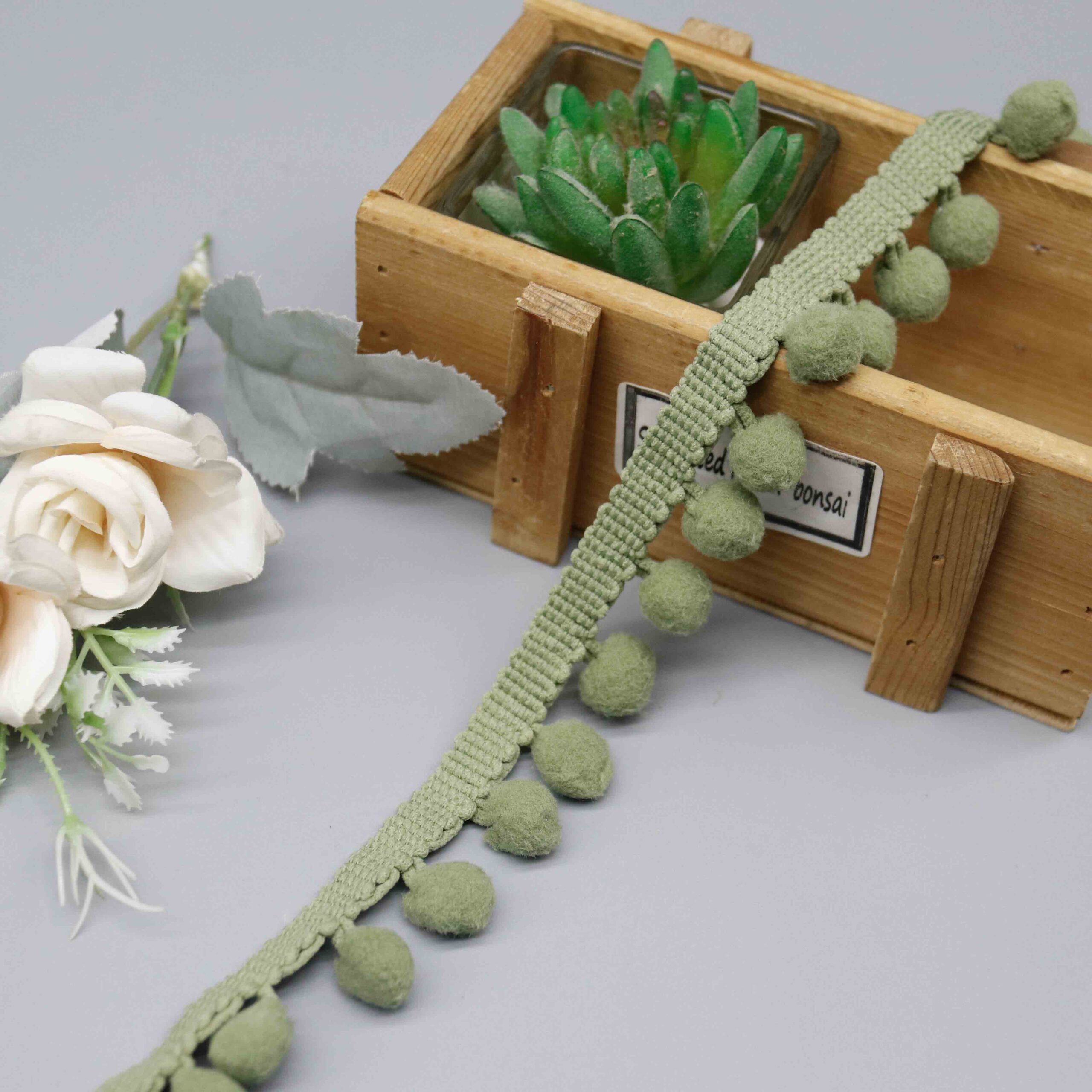 Front-facing shot of Olive Fuzzy Nylon Pompom Lace Trim, showing sage green pompom balls and braided bobble header clearly.