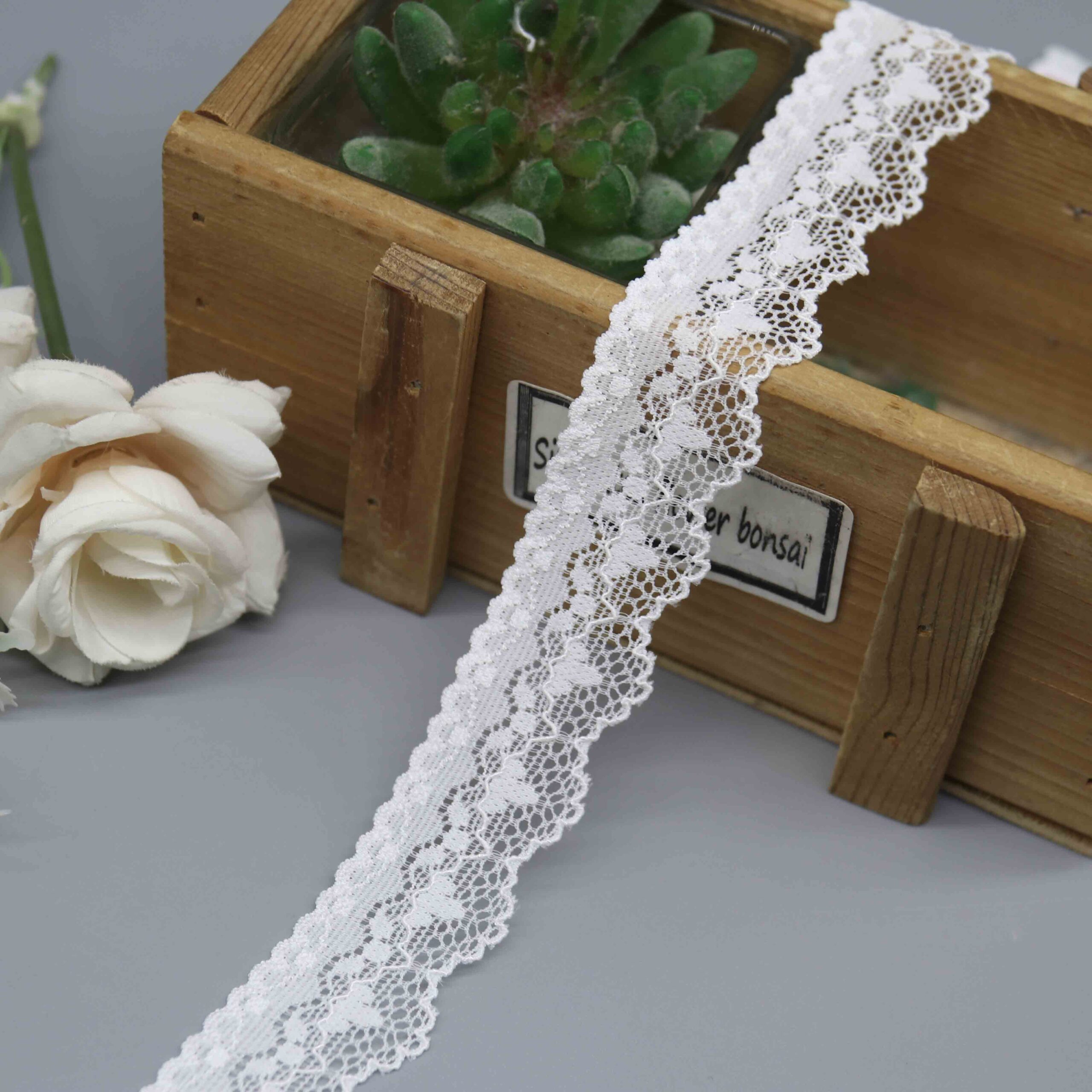 A close-up studio shot of a 2.5cm wide narrow stretch lace in white, showing its delicate floral pattern and symmetrical scalloped edges.