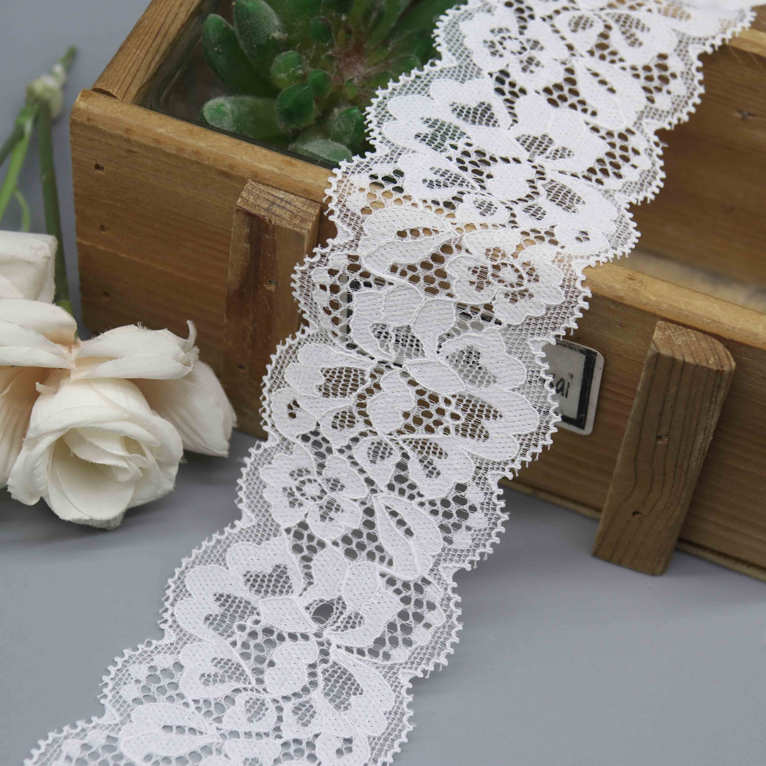 Studio photo of a 5cm wide white stretch galloon lace, featuring a symmetrical floral scroll pattern and double scalloped edges