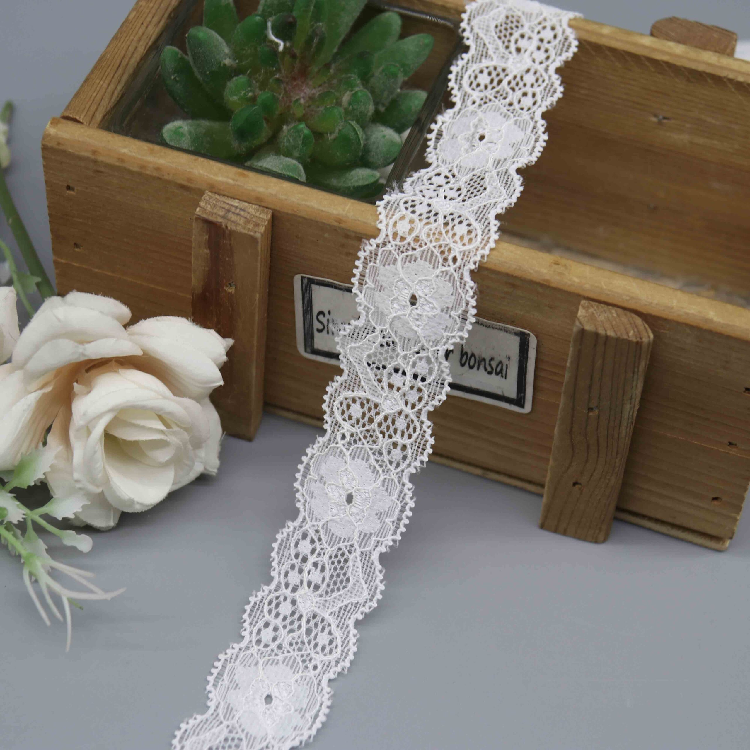 A studio photo of a 2.5cm wide white floral stretch lace trim, featuring a pattern of repeating floral medallions and scalloped edges.