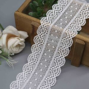 A studio shot of a 6cm wide, white stretch galloon lace with a geometric pattern and scalloped edges, displayed in a decorative wooden box