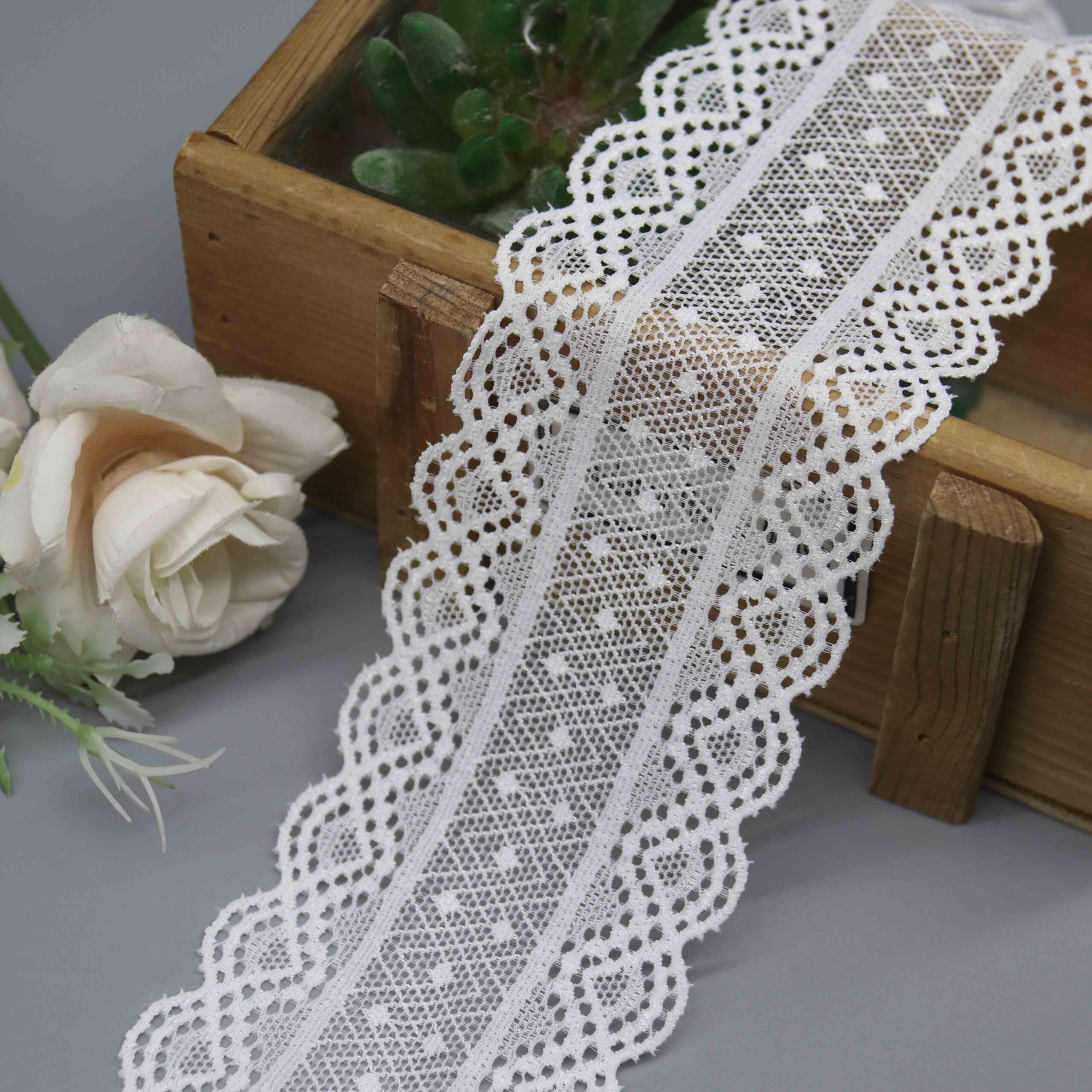 A studio shot of a 6cm wide, white stretch galloon lace with a geometric pattern and scalloped edges, displayed in a decorative wooden box
