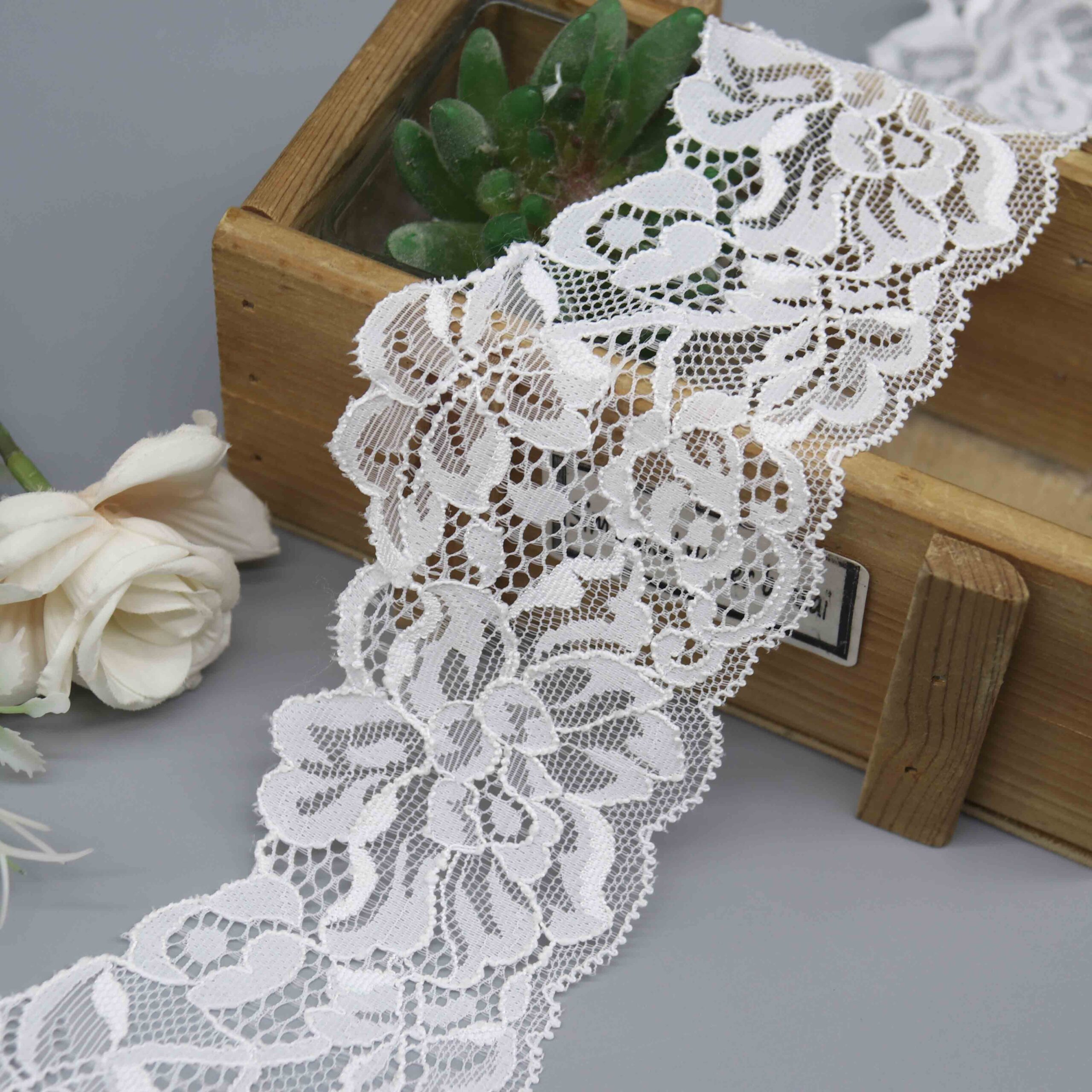 A product image of a 7cm wide white floral stretch lace, highlighting its intricate corded detail and scalloped edges.