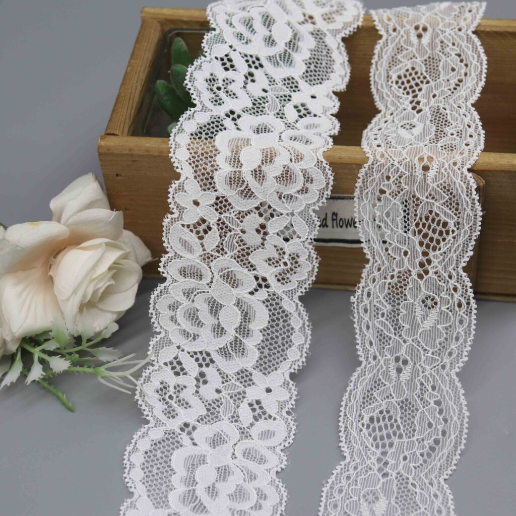 Two widths of a white floral stretch lace trim with scalloped edges, displayed in a wooden box with a decorative flower