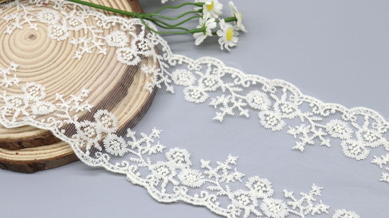 A close-up of a 10.5cm wide off-white polyester embroidered tulle lace with a symmetrical floral pattern and scalloped edges.