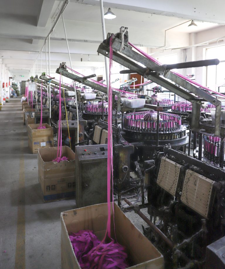 Row of industrial lace making machines producing vibrant pink lace trims in a factory setting.