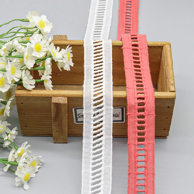 Close-up of cotton embroidered lace trim in white and pink colors, displayed in a wooden box with decorative white flowers, ideal for garment and craft embellishments.