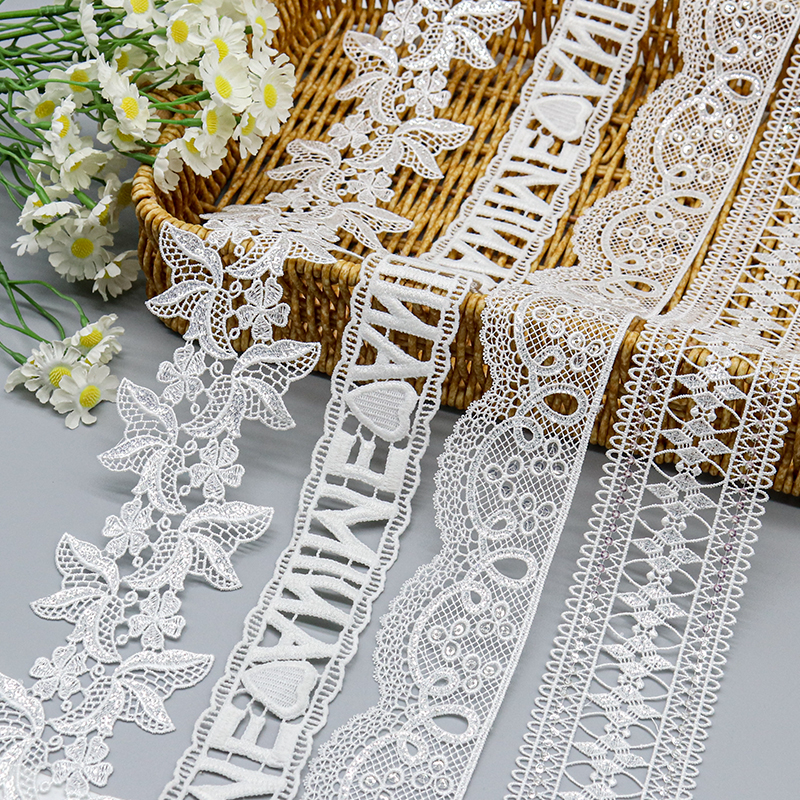 Assorted white water soluble lace trims featuring intricate floral, script, and geometric embroidery patterns displayed on a woven basket.