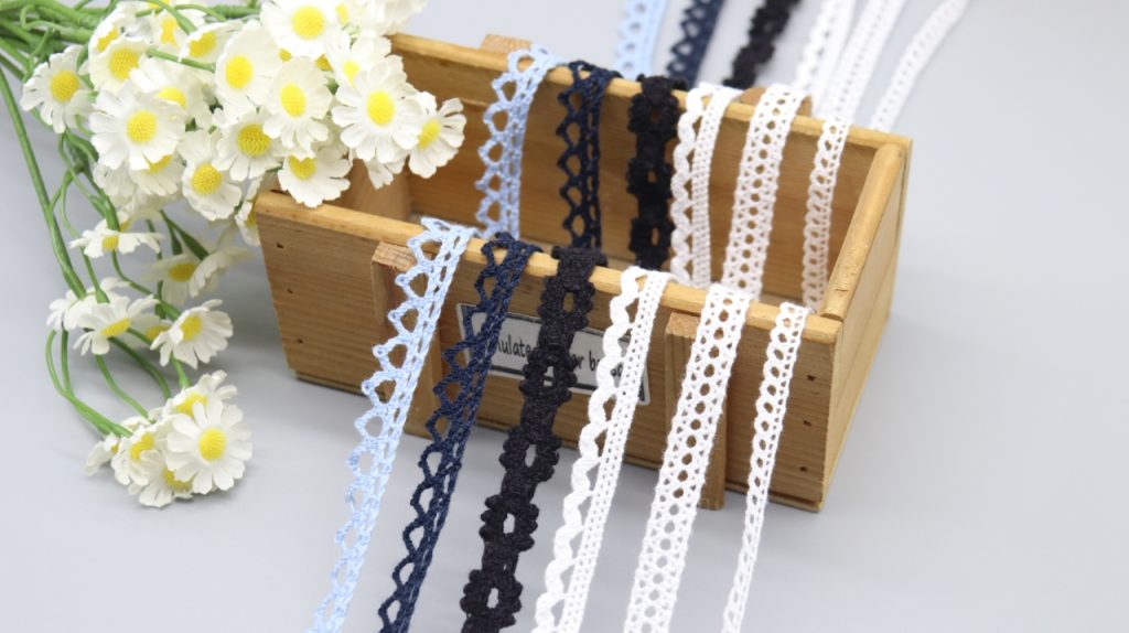 Assorted cotton thread lace trims in pastel blue, navy blue, black, white, and beige, displayed on a wooden box with daisies.