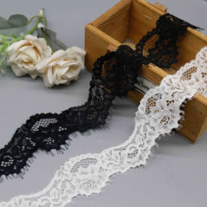 Close-up of elegant French stretch lace trim ribbon in black and white, soft and elastic with intricate floral vine patterns of flowers and leaves on a sheer mesh base; scalloped wave edges on both sides feature delicate eyelash fringe for a romantic, lightweight flutter, ideal for ladies' lingerie and neckline accents.